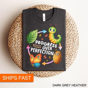 May include: Dark grey heather t-shirt with a graphic design of a caterpillar, butterfly, and a ladybug. The text "Progress over Perfection" is printed on the shirt.