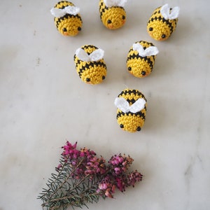 May include: Six crocheted bumblebees with black and yellow stripes and white wings. The bees are arranged in a circle on a white marble surface. A small sprig of pink heather is in the bottom center of the image.