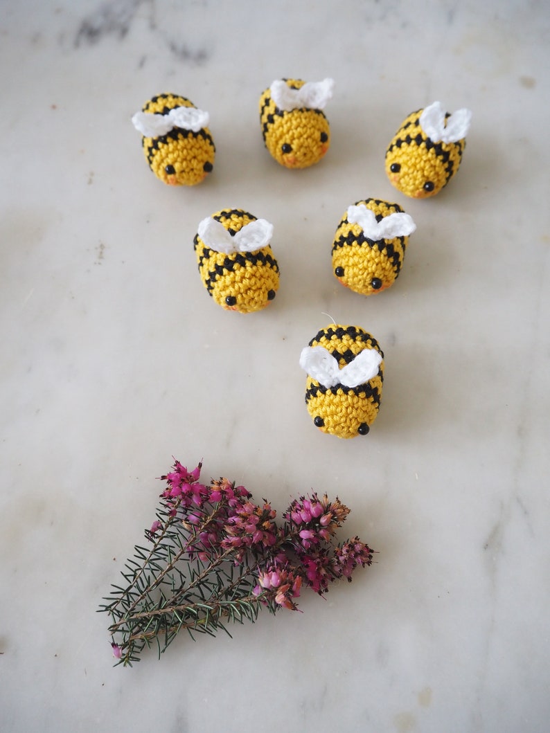 May include: Six crocheted bumblebees with black and yellow stripes and white wings. The bees are arranged in a circle on a white marble surface. A small sprig of pink heather is in the bottom center of the image.