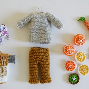 Peut inclure: Un ensemble d'articles au crochet comprenant un pull gris, un pantalon marron, un gilet blanc et marron, une écharpe florale et une variété de fruits au crochet, notamment des oranges et des carottes.