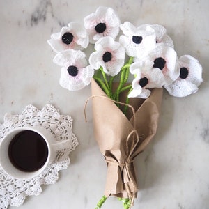 May include: A bouquet of white crocheted flowers with black centers, wrapped in brown paper and tied with twine. The flowers are arranged in a vase and are sitting on a white marble surface.