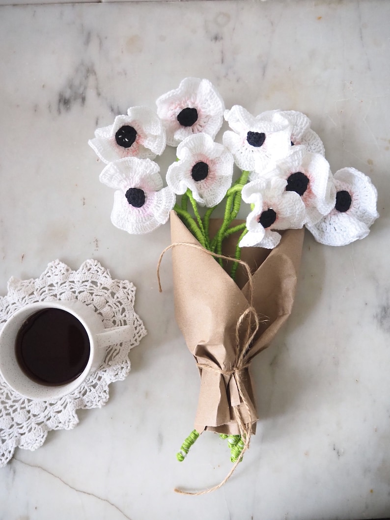 May include: A bouquet of white crocheted flowers with black centers, wrapped in brown paper and tied with twine. The flowers are arranged in a vase and are sitting on a white marble surface.