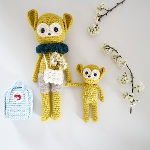 May include: Two crocheted stuffed animals, one larger and one smaller, both yellow with brown accents. The larger one is wearing a teal collar and holding a white crocheted bag. The smaller one is holding the larger one's hand. Both are on a white background with a branch of white flowers.