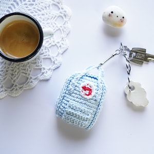 Peut inclure: Un porte-clés en forme de sac à dos bleu clair crocheté avec un logo rouge. Le porte-clés est attaché à un anneau de clé argenté avec une ficelle noire et blanche.