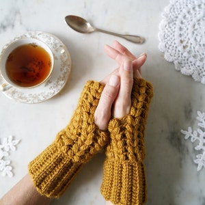 Peut inclure: Une paire de mitaines sans doigts en crochet jaune moutarde avec un motif de point texturé. Les mitaines sont portées par une personne avec les mains jointes.