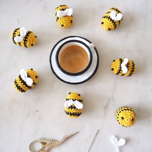 May include: A set of seven crocheted bumblebees in yellow and black with white wings, arranged around a white saucer with a cup of espresso. A pair of gold scissors and a needle are also included.