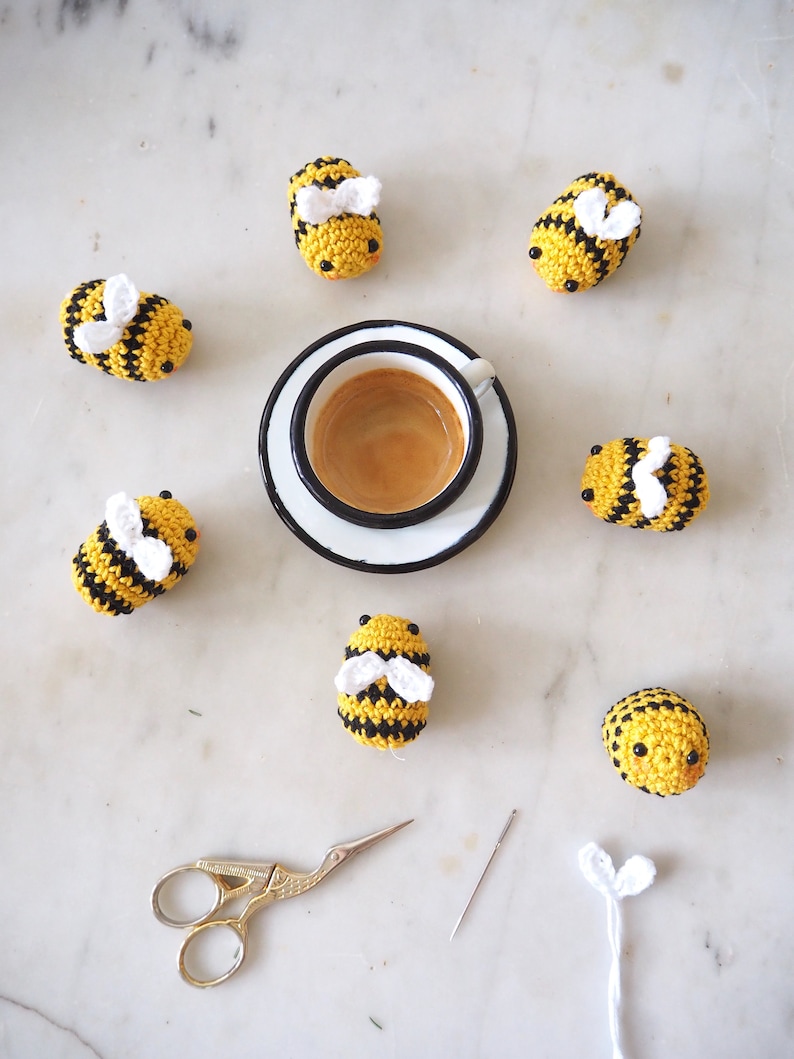 May include: A set of seven crocheted bumblebees in yellow and black with white wings, arranged around a white saucer with a cup of espresso. A pair of gold scissors and a needle are also included.