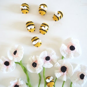 May include: A bouquet of white crocheted flowers with black centers and green stems. Seven crocheted bumblebees are scattered around the flowers. The bumblebees are yellow with black stripes and white wings.