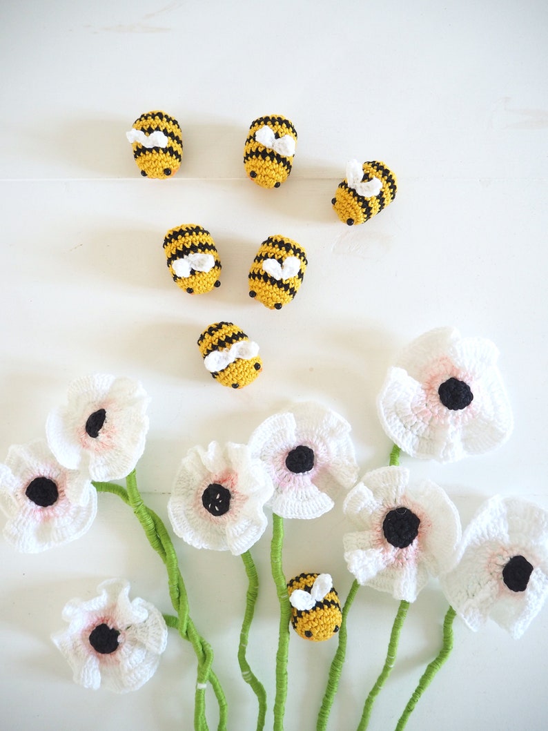 May include: A bouquet of white crocheted flowers with black centers and green stems. Seven crocheted bumblebees are scattered around the flowers. The bumblebees are yellow with black stripes and white wings.