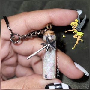 Könnte beinhalten: Eine durchsichtige Glasflasche gefüllt mit schimmerndem Glitzer und einem silbernen sternförmigen Zauberstab-Anhänger. Die Flasche ist an einem silbernen Schlüsselanhänger befestigt und hat einen Korkverschluss. Die Flasche wird in einer Hand mit weißem Nagellack gehalten. Das Bild zeigt auch eine Zeichnung von Tinkerbell mit einem Zauberstab.