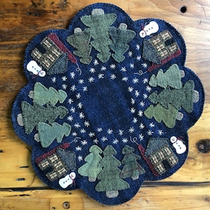 May include: A blue felt Christmas tree ornament with a scalloped edge. The ornament features a winter scene with green trees, white snowmen, and brown houses. The background is a dark blue felt with white embroidered snowflakes.