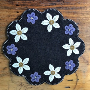 May include: A round, dark blue felt coaster with a scalloped edge. The coaster is decorated with white and yellow flowers and purple flowers.