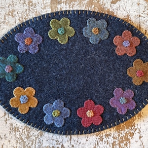May include: An oval-shaped, navy blue felt placemat decorated with colorful felt flowers. The flowers are in shades of orange, pink, purple, blue, and green, each with a contrasting center. The edge is stitched with a light brown thread.
