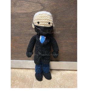 May include: Crocheted doll wearing a black face mask, a black suit, a blue tie, and blue pants. The doll has a light brown complexion and black eyes. The doll is standing on a wooden surface.