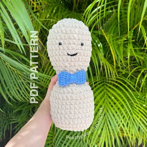 Puede incluir: Un juguete de peluche tejido a crochet con forma de persona con una pajarita azul. El juguete es beige y tiene una cara sonriente con ojos negros. El juguete se está sosteniendo frente a un fondo verde frondoso.
