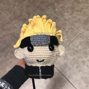 Könnte beinhalten: Gehäkelte Golf- Schlägerkopf-Hülle mit einem Comic-Charakter mit gelben Haaren, einem schwarzen Stirnband und einem schwarzen und grauen Stirnschutz. Der Charakter hat ein lächelndes Gesicht mit schwarzen Augen und rosa Wangen. Der Text "PeachyMeechesCrochet" ist auf der Hülle sichtbar.