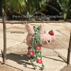 May include: A pink crocheted pig hanging from a wooden stick with green and pink crocheted flowers and leaves. The pig has a red crocheted nose.