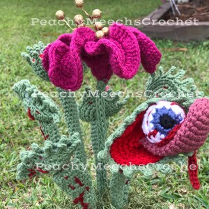 May include: A crocheted plant with a pink flower and a red and white eye. The plant is green and has a textured surface. The eye is open and has a blue iris and a black pupil.