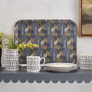 Pode incluir: Uma bandeja cinza e branca com padrão floral e borda dourada, quatro canecas brancas com padrão floral preto e dourado, um jarro branco com padrão floral preto e dourado e um conjunto de pratos brancos com borda dourada.