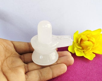 May include: White selenite Shiva lingam statue, a Hindu deity, on a hand with a yellow flower in the background.