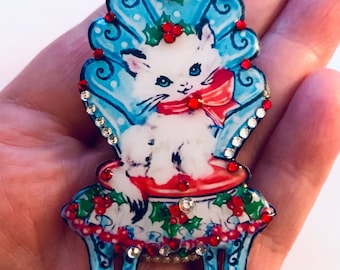 Puede incluir: Un gatito blanco con un lazo rojo se sienta en una silla azul y blanca con acebo rojo y bayas. La silla está decorada con diamantes de imitación.