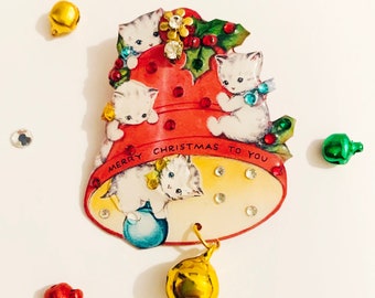 Puede incluir: Un adorno navideño vintage con forma de campana con cuatro gatitos y el texto "Merry Christmas to You". La campana es roja y blanca con ribetes dorados y tiene una pequeña campana dorada colgando de la parte inferior.