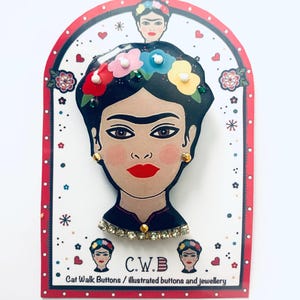 May include: A colourful illustrated brooch featuring Frida Kahlo with a flower crown and rhinestones. The brooch is on a white background with a red border and the text "C.W.B. Cat Walk Buttons / illustrated buttons and jewellery".