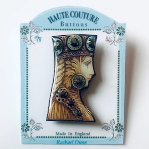May include: A brown and gold button with a detailed image of a woman's face wearing a crown. The button is decorated with rhinestones and has the text "HAUTE COUTURE Buttons" and "Made in England Rachel Dunn" on the card.