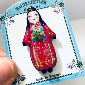 Könnte beinhalten: Vintage Brosche in Puppenform mit rotem Kleid, goldenen Details und einem türkisfarbenen und goldenen Mittelstück. Die Brosche ist auf einer blauen Karte mit dem Text "HAUTE COUTURE" und "Made in England" befestigt.