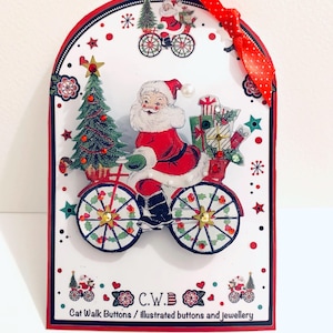 May include: A white Christmas card with a red border and a red ribbon. The card features a vintage illustration of Santa Claus riding a bicycle with a Christmas tree and presents. The text on the card reads "C.W.B. Cat Walk Buttons / illustrated buttons and jewellery."
