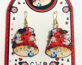 Puede incluir: Un par de pendientes de campana de Navidad rojos y blancos con un diseño de gatitos y pedrería. Los pendientes están en una tarjeta que dice "C.W.B. Cat Walk Buttons / illustrated buttons and jewellery".