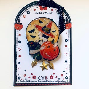 May include: A Halloween-themed brooch featuring a woman in a witch's hat holding a jack-o'-lantern. The brooch is made of metal and has a gold finish. The woman is wearing a black dress and a green scarf. The brooch is decorated with rhinestones and stars. The text "HALLOWEEN" and "C.W.B. Cat Walk Buttons / illustrated buttons and jewellery" is printed on the card.