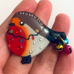 May include: A red and white robin Christmas ornament with a black and white polka dot bow and the words "HO HO HO" on the back. The ornament has a string of lights and jingle bells attached.
