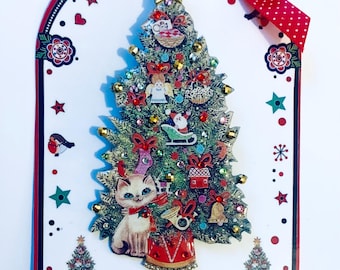 Puede incluir: Un botón con forma de árbol de Navidad con una figura de gato, decorado con pedrería, adornos y una estrella en la parte superior. El botón está rodeado por un borde rojo, blanco y negro con una cinta roja con lunares blancos. El texto "C.W.B Cat Walk Buttons / illustrated buttons and jewellery" está impreso en la parte inferior.
