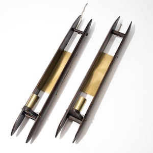 May include: Two elongated, dark brown wooden weaving shuttles featuring pointed ends and a central brass plate. Each shuttle includes a cylindrical brass bobbin holder positioned near one end, with a small metal chain attached to the top.