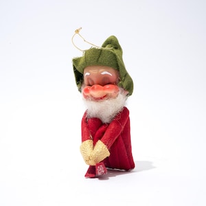 Pode incluir: Um enfeite de Natal vintage de um duende sorridente. O duende tem um rosto rosado, barba branca e usa um chapéu verde, casaco vermelho e luvas douradas. Uma corda dourada é presa ao chapéu para pendurar.