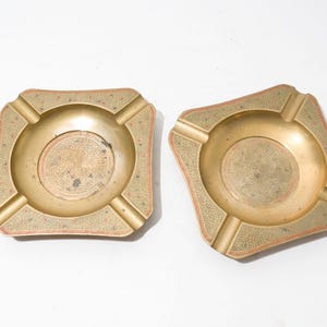 May include: Two gold-colored metal ashtrays with intricate designs. Each ashtray has four indentations for cigarettes and a circular design in the center.