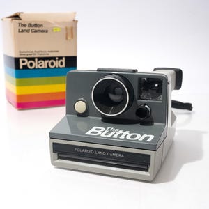 May include: A gray Polaroid "The Button" Land Camera with a black lens and flash. The camera has a white button and the name "The Button" printed on the front. A colorful box with the Polaroid logo is in the background.
