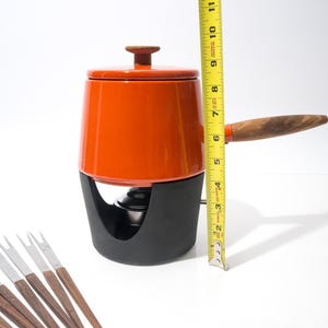 May include: An orange fondue pot with a black base and wooden handle, measuring approximately 10 inches tall. Includes a lid with a wooden knob and several fondue forks with wooden handles.