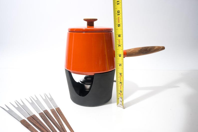 May include: An orange fondue pot with a black base and wooden handle, measuring approximately 10 inches tall. Includes a lid with a wooden knob and several fondue forks with wooden handles.