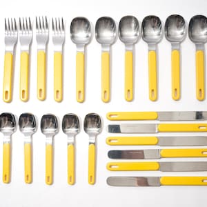 Puede incluir: Un juego de cubiertos vintage de acero inoxidable con mangos amarillos. El juego incluye tenedores, cucharas y cuchillos de mantequilla. Los utensilios están dispuestos sobre una superficie blanca.