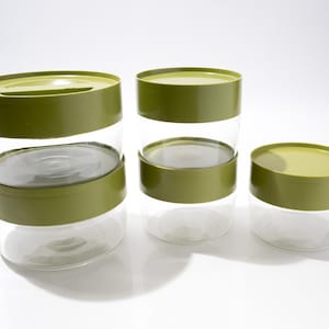 May include: A set of five clear glass storage containers with olive green lids. The containers are arranged in three stacks of varying heights, featuring cylindrical bodies and flat, solid-coloured lids that fit securely onto the glass bases.