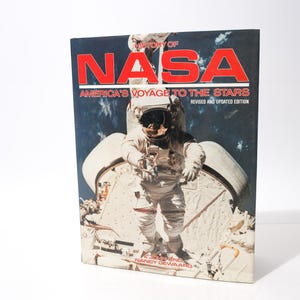 May include: A book titled "History of NASA: America's Voyage to the Stars" with a revised and updated edition. The cover features a large red "NASA" logo and an astronaut in a spacesuit. The background is a dark blue space scene.