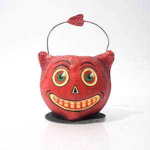 May include: A red devil-faced Halloween candy pail with a black handle and a small red glittered heart on top. The pail has green and yellow eyes, a black triangle nose, and a yellow and red smiling mouth.