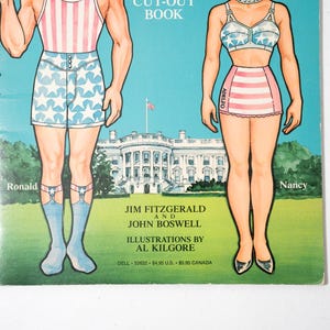 May include: Vintage paper doll book cover featuring illustrations of Ronald and Nancy in patriotic attire. The background shows the White House and a blue sky. The book is titled "Cut-Out Book" and includes the names of the authors and illustrator.