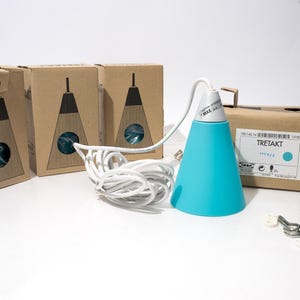 May include: A bright turquoise cone-shaped pendant lamp with a white cord and plug, accompanied by three brown cardboard boxes labeled TRETAKT. The lamp features a white top section with text reading MAX 25W, and includes small white plastic hardware pieces.