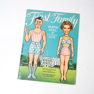 May include: A vintage "First Family" paper doll and cut-out book featuring illustrations of Ronald and Nancy Reagan. The cover is turquoise with the title in white script and illustrations of the couple in swimwear.