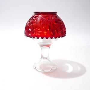 Pode incluir: Um castiçal de vidro vermelho com um padrão floral decorativo na sombra. A sombra assenta sobre uma haste e base de vidro transparente. O castiçal foi concebido para conter uma pequena vela.