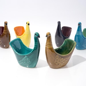 May include: A collection of ceramic bird-shaped vases in various colors, including brown, yellow, teal, and blue. Each vase has a unique design, with some featuring textured patterns. The vases are arranged on a white surface.