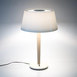 May include: A white table lamp with a conical lampshade. The lamp has a cream-coloured stem and a circular base. The lampshade is white and has a dark circular design at the top. The lamp is on a white surface.